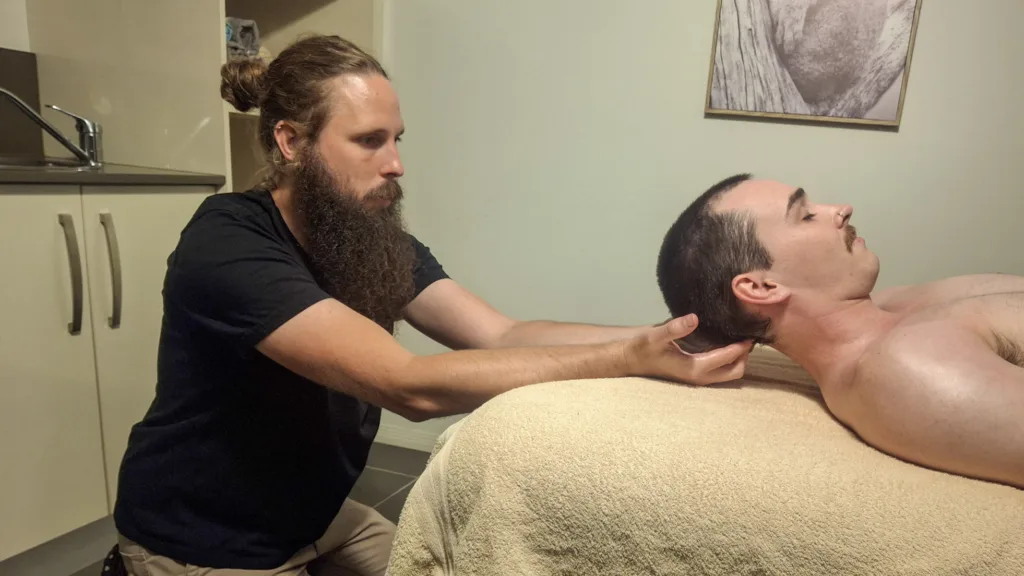 A Massage Therapist Performing A Head Massage On A Male Client Lying On A Couch In A Contemporary Clinic, Aimed At Relieving Migraine Discomfort.