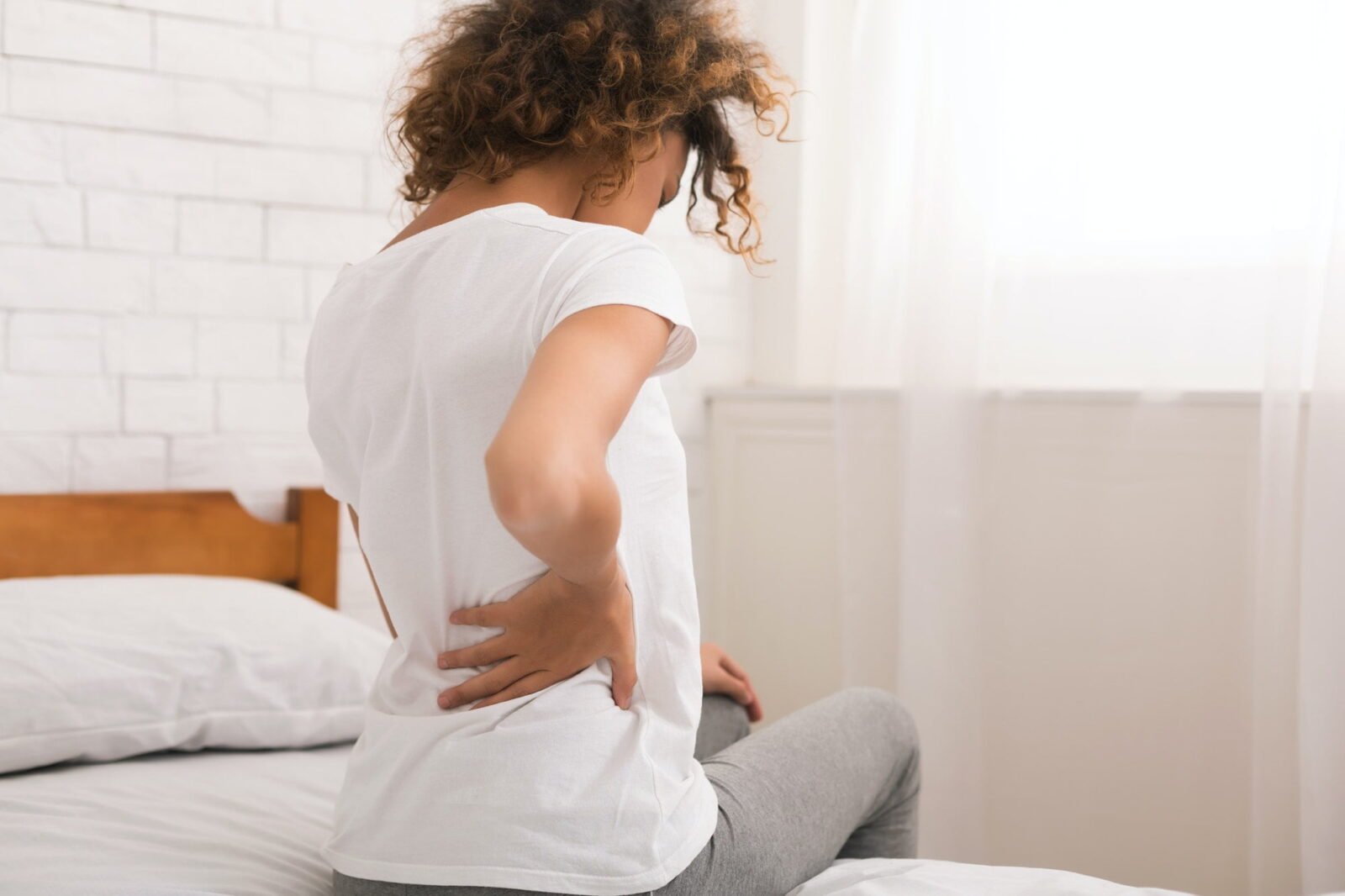 A woman sitting on the edge of a bed, holding her lower back in discomfort, indicating symptoms of back pain, sciatica, and nerve pain, seeking help from a Back Pain Specialist.