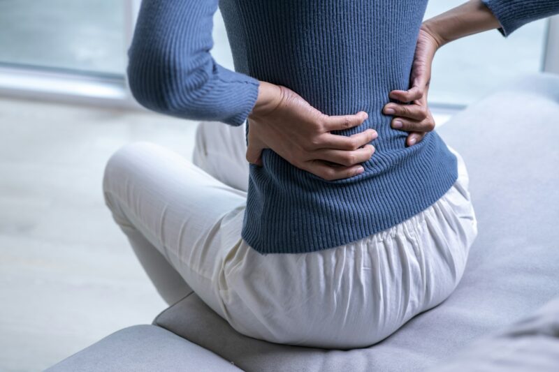 A woman sitting on a couch, holding her lower back with both hands, depicting discomfort and the need for back pain massage.