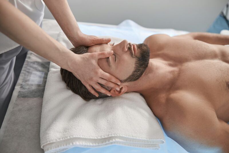 Fullon Life: Massage Expert In Zillmere, Qld 3 A Massage Therapist Working On The Head Of A Male Client Lying On A Couch In A Contemporary Clinic, Emphasizing Services Covered Under Massage Health Fund Rebate Programs.