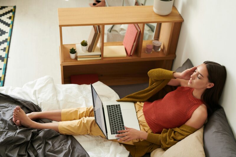 A woman sitting on a bed with a laptop, holding her head and appearing distressed due to migraines.