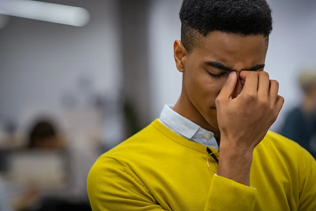 Remedial Massage For Long-Term Migraine Management 1 A Young Man Touching The Bridge Of His Nose With A Pained Expression, Showing Discomfort From A Migraine.